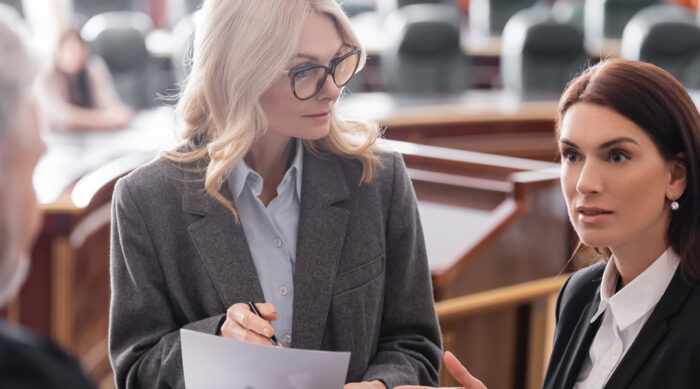 Two female defamation attorneys review lawsuit documents in court with a male judge.
